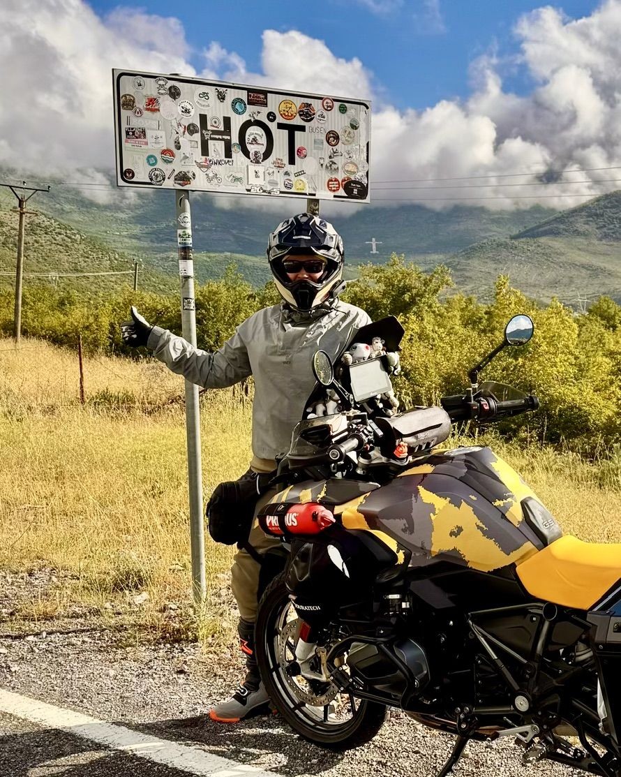 Angela on her motorbike next to a sticker-covered village sign that reads HOT.