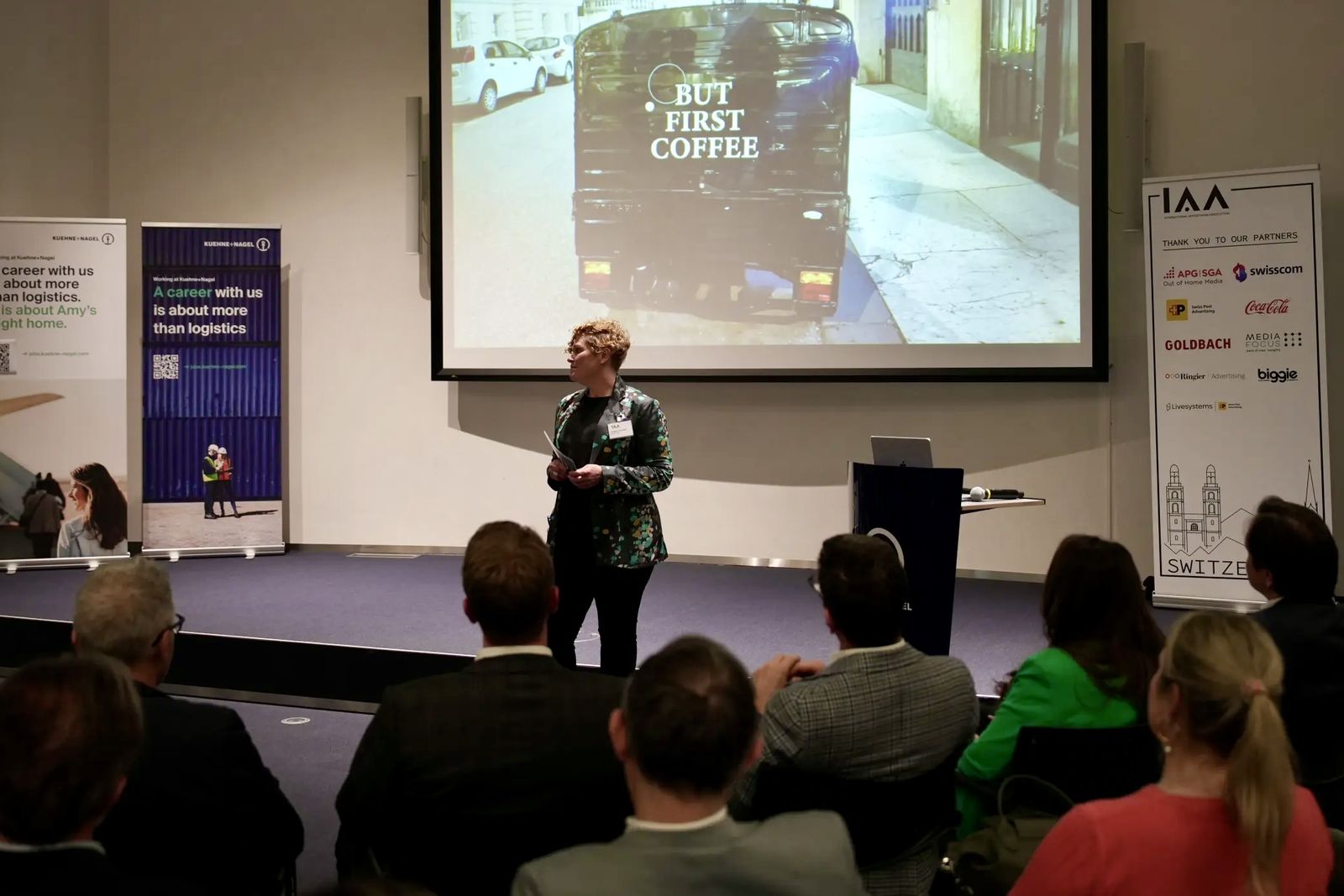 Angela speaking on stage in front of an audience. The slide behind her shows a van with the words But first coffee.